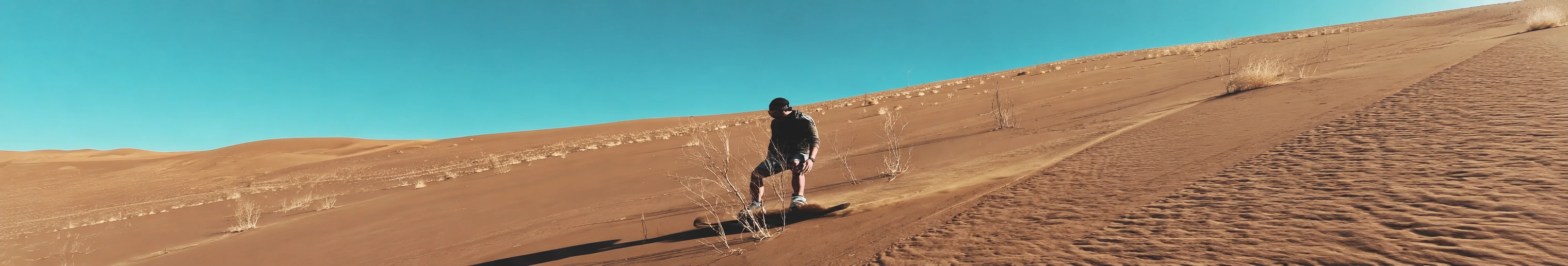 A sandboarder riding a sunlit desert dune beneath a turquoise sky