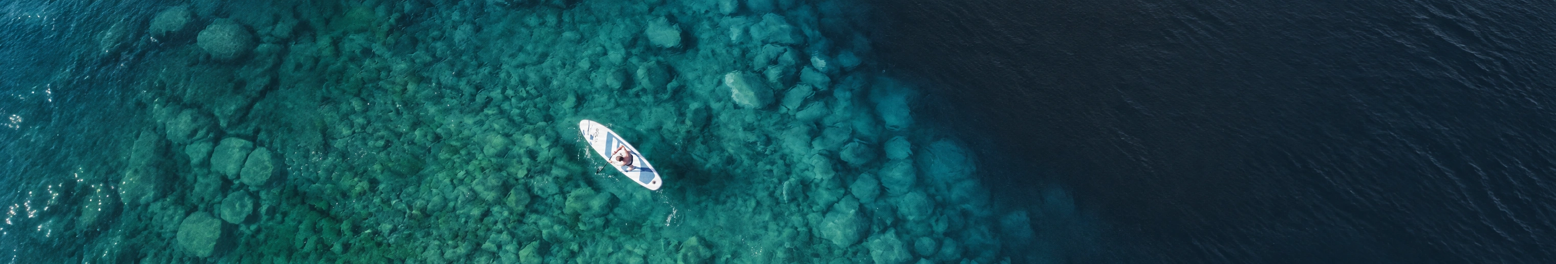 A paddleboarder above a rocky turquoise seabed beside deeper blue water at Pine Bay